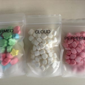 May include: Three clear plastic bags filled with colorful marshmallows. The bags are labeled "CHARMED," "CLOUD," and "PEPPERMINT." The "CHARMED" bag contains green, blue, pink, and yellow marshmallows. The "CLOUD" bag has white marshmallows, and the "PEPPERMINT" bag has pink marshmallows.
