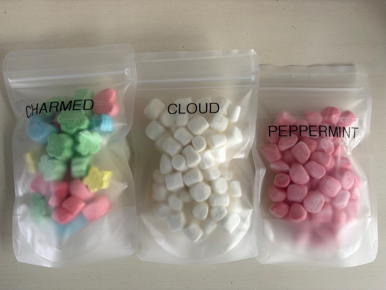 May include: Three clear plastic bags filled with colorful marshmallows. The bags are labeled "CHARMED," "CLOUD," and "PEPPERMINT." The "CHARMED" bag contains green, blue, pink, and yellow marshmallows. The "CLOUD" bag has white marshmallows, and the "PEPPERMINT" bag has pink marshmallows.