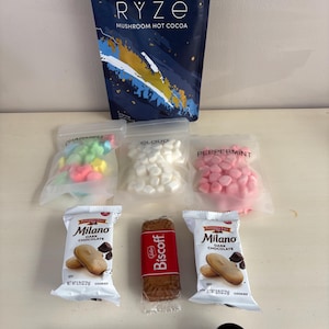 May include: An assortment of treats including a bag of RYZE mushroom hot cocoa, bags of marshmallows and candies, Milano dark chocolate cookies, a Biscoff cookie, and a black scoop. The hot cocoa bag is navy blue with white text.