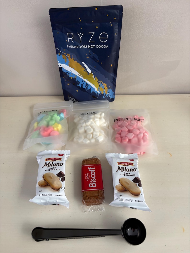 May include: An assortment of treats including a bag of RYZE mushroom hot cocoa, bags of marshmallows and candies, Milano dark chocolate cookies, a Biscoff cookie, and a black scoop. The hot cocoa bag is navy blue with white text.