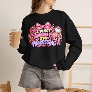 May include: A black long-sleeve sweatshirt featuring a graphic print with the text I'M NOT IGNORING YOU, I'M PROCESSING. The design includes illustrations of pink bows, lipstick, a compact mirror, and small hearts in shades of pink and purple.