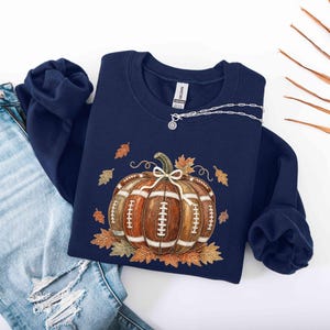 May include: A navy blue crewneck sweatshirt featuring a graphic of a brown football-textured pumpkin with white laces and a small bow on top, surrounded by scattered orange and brown autumn leaves, paired with light blue distressed denim jeans.