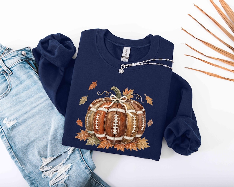 May include: A navy blue crewneck sweatshirt featuring a graphic of a brown football-textured pumpkin with white laces and a small bow on top, surrounded by scattered orange and brown autumn leaves, paired with light blue distressed denim jeans.