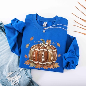 May include: A royal blue long-sleeve sweatshirt featuring a central graphic of a brown pumpkin designed with the texture and white laces of an American football. The pumpkin is surrounded by scattered orange and brown autumn leaves.