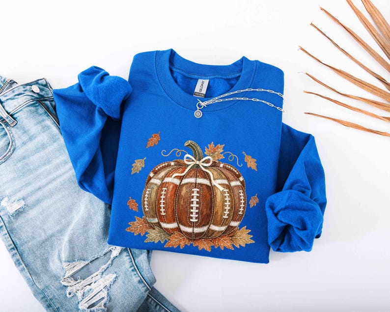 May include: A royal blue long-sleeve sweatshirt featuring a central graphic of a brown pumpkin designed with the texture and white laces of an American football. The pumpkin is surrounded by scattered orange and brown autumn leaves.