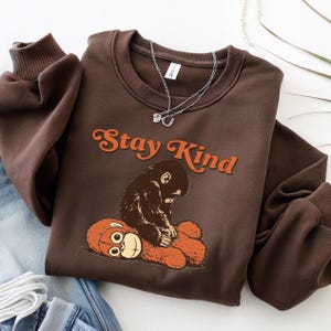 May include: A brown sweatshirt with the text "Stay Kind" in orange script. The graphic features a monkey hugging a teddy bear. The sweatshirt has a crew neck and long sleeves with ribbed cuffs. A silver necklace is draped over the sweatshirt.