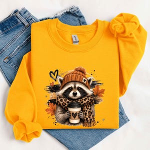 Pode incluir: Uma camisola de gola redonda em amarelo vivo com um gr&aacute;fico de um guaxinim a usar um gorro de malha castanho e um cachecol com padr&atilde;o de leopardo, segurando um copo de caf&eacute; com um cora&ccedil;&atilde;o, rodeado por folhas de outono e tra&ccedil;os pretos.