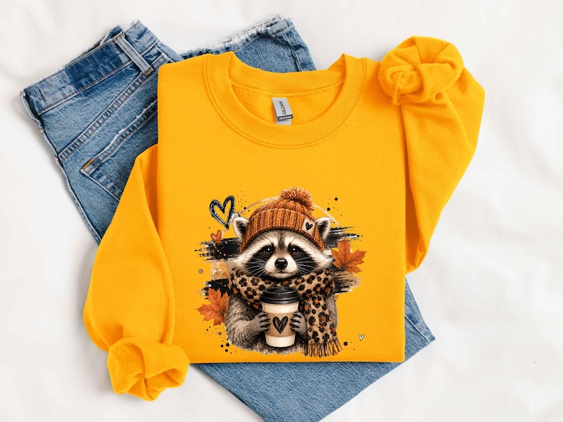 Pode incluir: Uma camisola de gola redonda em amarelo vivo com um gr&aacute;fico de um guaxinim a usar um gorro de malha castanho e um cachecol com padr&atilde;o de leopardo, segurando um copo de caf&eacute; com um cora&ccedil;&atilde;o, rodeado por folhas de outono e tra&ccedil;os pretos.