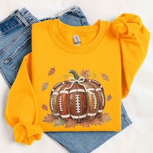 May include: A bright yellow crewneck sweatshirt featuring a central graphic of a pumpkin designed to look like a brown leather football with white laces. The pumpkin is accented with a small white bow and surrounded by scattered autumn leaves.