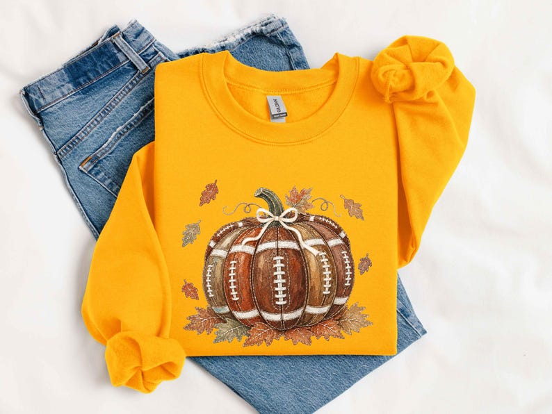 May include: A bright yellow crewneck sweatshirt featuring a central graphic of a pumpkin designed to look like a brown leather football with white laces. The pumpkin is accented with a small white bow and surrounded by scattered autumn leaves.