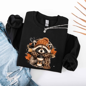 Pode incluir: Uma camisola preta de manga comprida com o gr&aacute;fico de um guaxinim a usar um gorro de malha laranja e um cachecol com padr&atilde;o de leopardo, segurando uma ch&aacute;vena de caf&eacute; com um cora&ccedil;&atilde;o, rodeado por folhas de outono laranja e um pequeno cora&ccedil;&atilde;o preto.