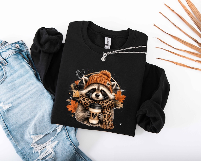 Pode incluir: Uma camisola preta de manga comprida com o gr&aacute;fico de um guaxinim a usar um gorro de malha laranja e um cachecol com padr&atilde;o de leopardo, segurando uma ch&aacute;vena de caf&eacute; com um cora&ccedil;&atilde;o, rodeado por folhas de outono laranja e um pequeno cora&ccedil;&atilde;o preto.