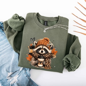 Pode incluir: Uma camisola de gola redonda verde-azeitona com um gr&aacute;fico de um guaxinim a usar um gorro de malha laranja e um cachecol com padr&atilde;o de leopardo. O guaxinim segura um copo de caf&eacute; com um cora&ccedil;&atilde;o, rodeado por folhas de outono cor de laranja.