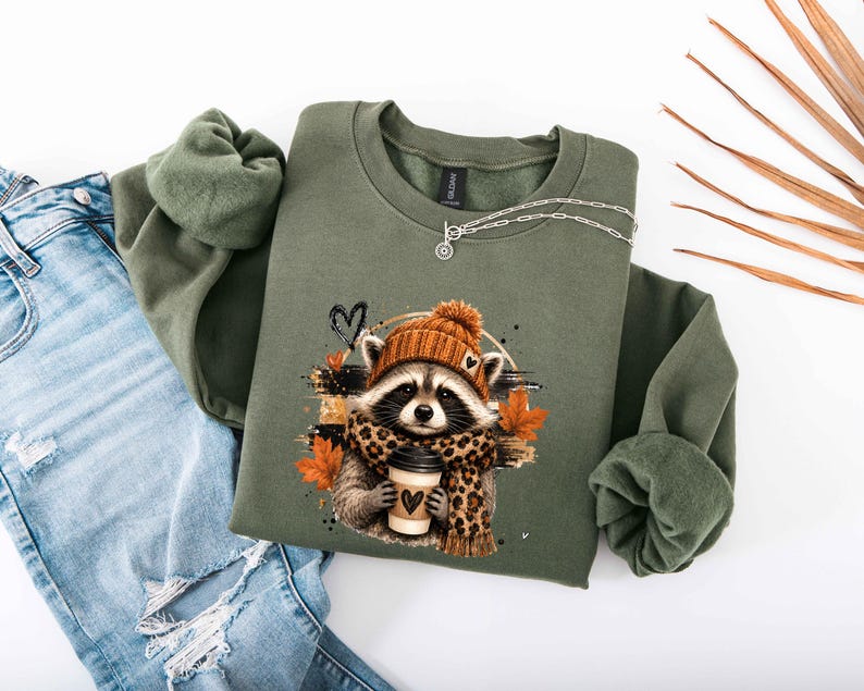 Pode incluir: Uma camisola de gola redonda verde-azeitona com um gr&aacute;fico de um guaxinim a usar um gorro de malha laranja e um cachecol com padr&atilde;o de leopardo. O guaxinim segura um copo de caf&eacute; com um cora&ccedil;&atilde;o, rodeado por folhas de outono cor de laranja.