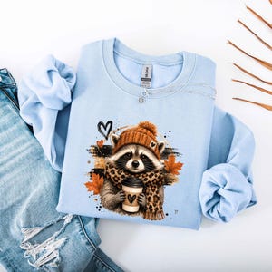 Pode incluir: Uma camisola de gola redonda azul-clara com um gr&aacute;fico de um guaxinim a usar um gorro de malha laranja e um cachecol com padr&atilde;o de leopardo, segurando uma ch&aacute;vena de caf&eacute;. O design inclui folhas de &aacute;cer cor de laranja e cora&ccedil;&otilde;es pretos.
