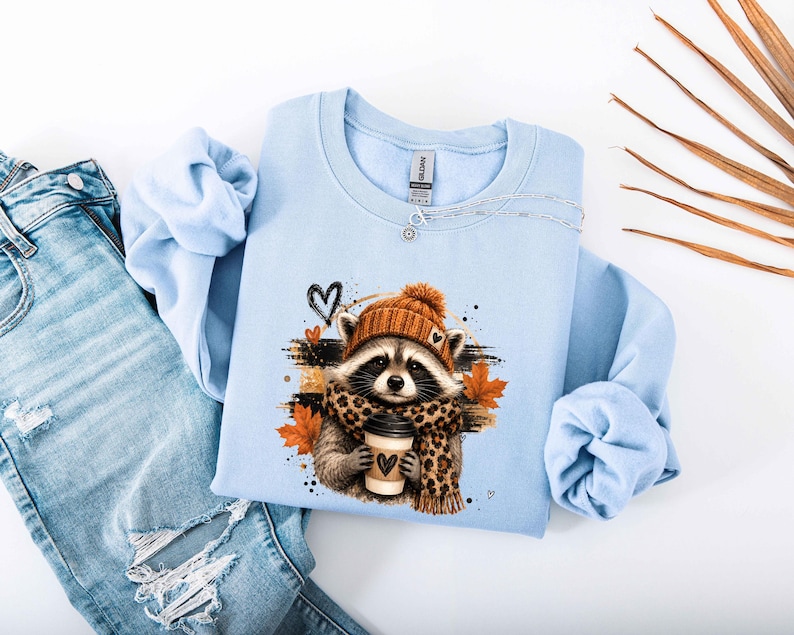 Pode incluir: Uma camisola de gola redonda azul-clara com um gr&aacute;fico de um guaxinim a usar um gorro de malha laranja e um cachecol com padr&atilde;o de leopardo, segurando uma ch&aacute;vena de caf&eacute;. O design inclui folhas de &aacute;cer cor de laranja e cora&ccedil;&otilde;es pretos.
