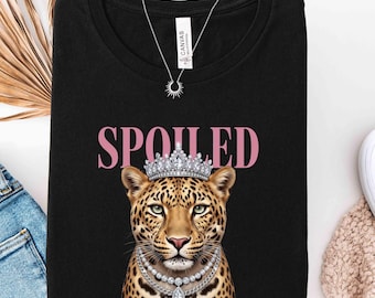 May include: Black t-shirt with the word "SPOILED" in pink letters above a leopard wearing a crown and a pearl necklace. A silver necklace hangs from the neckline. The shirt is folded.