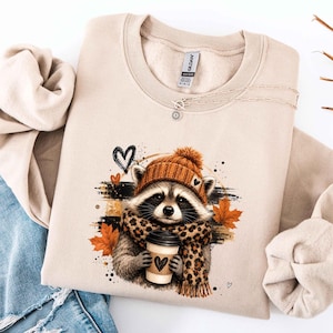 May include: A sand-colored crewneck sweatshirt featuring a graphic of a raccoon wearing an orange knit beanie and a leopard print scarf. The raccoon holds a coffee cup with a heart icon. Orange maple leaves and black hearts surround the raccoon.