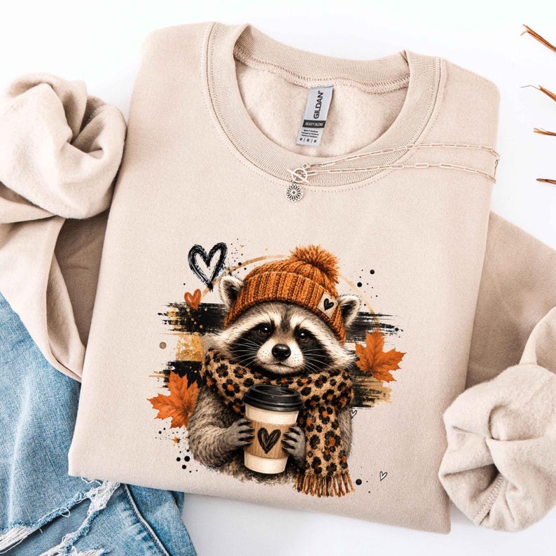 Pode incluir: Uma camisola de gola redonda cor de areia com um gr&aacute;fico de um guaxinim a usar um gorro de malha laranja e um cachecol com padr&atilde;o de leopardo. O guaxinim segura um copo de caf&eacute; com um cora&ccedil;&atilde;o. Folhas de &aacute;cer laranja e cora&ccedil;&otilde;es pretos.