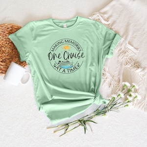Peut inclure: Un t-shirt &agrave; manches courtes vert clair avec un motif circulaire noir portant l'inscription "MAKING MEMORIES ONE CRUISE AT A TIME" entourant l'illustration d'un navire de croisi&egrave;re, d'un palmier et d'un petit soleil jaune.