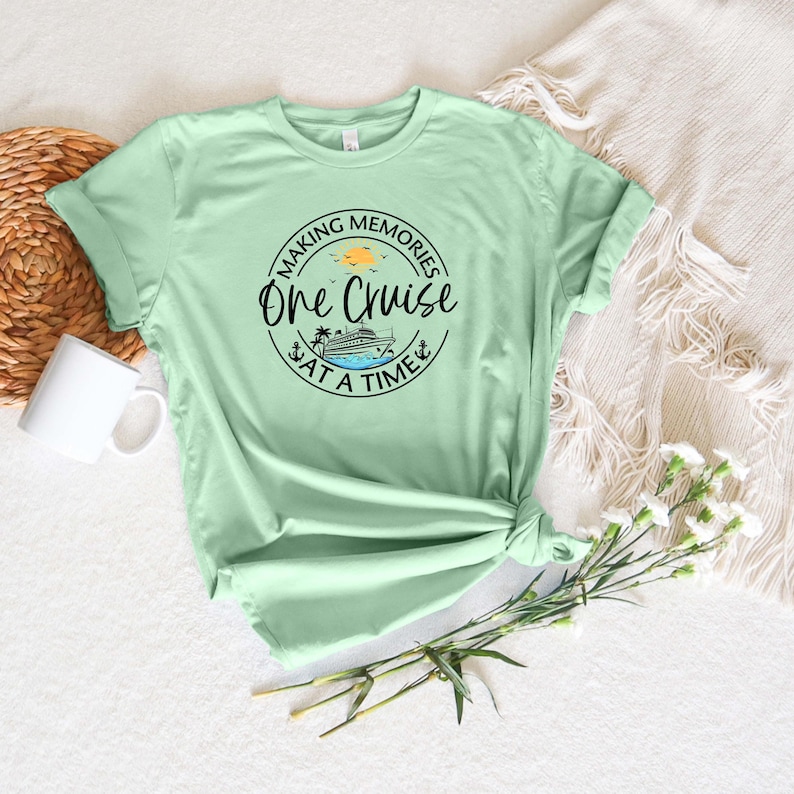 Peut inclure: Un t-shirt &agrave; manches courtes vert clair avec un motif circulaire noir portant l'inscription "MAKING MEMORIES ONE CRUISE AT A TIME" entourant l'illustration d'un navire de croisi&egrave;re, d'un palmier et d'un petit soleil jaune.