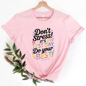 May include: A light pink t-shirt with the text "Don't Stress! It's Test Day Do your BEST!" in a colorful, playful font. The design includes various school-themed icons. The shirt is hanging on a wooden hanger.
