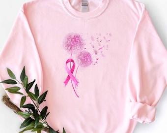 May include: A light pink crewneck sweatshirt featuring a graphic of two dandelion seed heads in shades of pink. One dandelion stem is wrapped in a pink awareness ribbon. The seeds appear to be blowing away toward the right side of the garment.