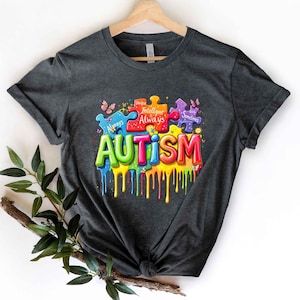 May include: Dark gray t-shirt with a colorful graphic design. The design features puzzle pieces and the word "AUTISM" in vibrant, dripping paint. The puzzle pieces have the words "Unique", "Intelligent", "Always", and "Sometimes".