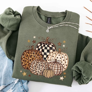 May include: An olive green crewneck sweatshirt featuring a graphic print of five pumpkins with various patterns including black and white checkered, leopard print, polka dots, and plaid, accented by small brown and cream stars and swirling vines.