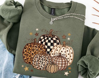 Pode incluir: Uma camisola de gola redonda verde-azeitona com uma impressão gráfica de cinco abóboras com vários padrões, incluindo xadrez preto e branco, estampado de leopardo, bolinhas e riscas, acentuada por pequenas estrelas castanhas e creme.