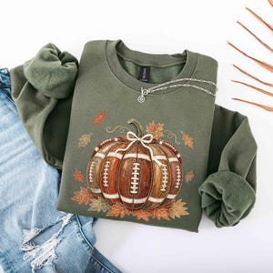 May include: An olive green crewneck sweatshirt featuring a graphic of a brown pumpkin designed with white football laces and a white bow on top, surrounded by scattered autumn leaves. The sweatshirt is paired with distressed blue denim jeans.