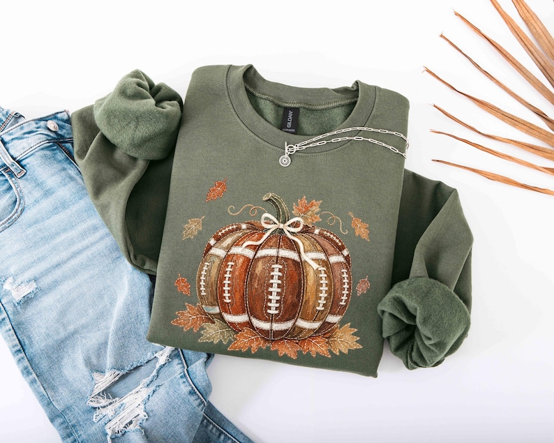 May include: An olive green crewneck sweatshirt featuring a graphic of a brown pumpkin designed with white football laces and a white bow on top, surrounded by scattered autumn leaves. The sweatshirt is paired with distressed blue denim jeans.