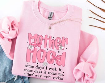 Pode incluir: Um moletom rosa claro com a palavra "Motherhood" em letras rosa com laços e flores. O texto "some days I rock it, some days it rocks me, either way we're rockin'" está impresso abaixo. Um colar de ouro está drapeado sobre o moletom.