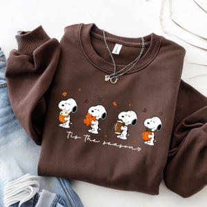 May include: A dark brown crewneck sweatshirt featuring a graphic print of the cartoon character Snoopy in four different poses with autumn-themed items including a drink, a leaf, a football, and a pumpkin, with the text 'Tis the season'.