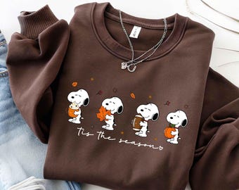 Peut inclure: Un sweat-shirt à col rond marron foncé avec une impression graphique du personnage de dessin animé Snoopy dans quatre poses différentes avec des éléments sur le thème de l'automne, incluant le texte 'Tis the season'.