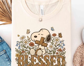 Peut inclure: T-shirt crème avec un dessin de Snoopy tenant une croix en bois, entouré de fleurs et de papillons. Le mot "BLESSED" est écrit en lettres marron fleuries. Le t-shirt est plié proprement.