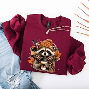 Pode incluir: Uma camisola bordeaux de manga comprida com o gr&aacute;fico de um guaxinim a usar um gorro de malha laranja e um cachecol com padr&atilde;o de leopardo. O guaxinim segura um copo de caf&eacute; com um &iacute;cone de cora&ccedil;&atilde;o. O design inclui folhas de outono laranja.