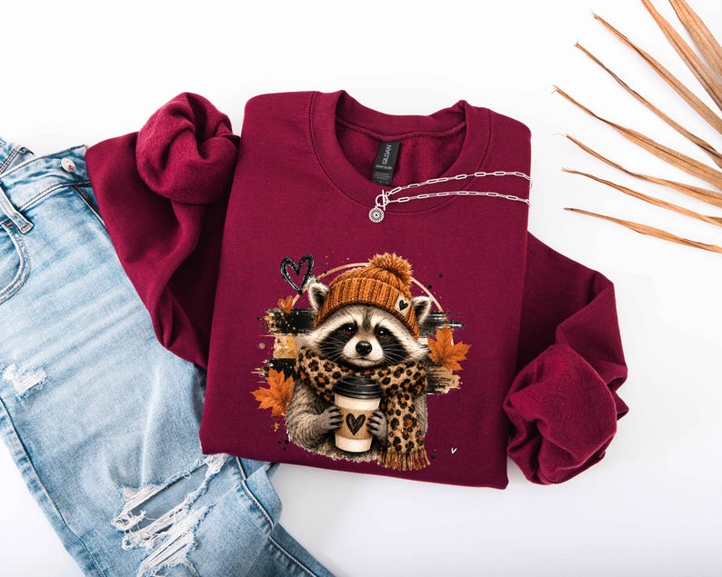 Pode incluir: Uma camisola bordeaux de manga comprida com o gr&aacute;fico de um guaxinim a usar um gorro de malha laranja e um cachecol com padr&atilde;o de leopardo. O guaxinim segura um copo de caf&eacute; com um &iacute;cone de cora&ccedil;&atilde;o. O design inclui folhas de outono laranja.