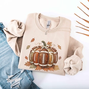 May include: A sand-colored crewneck sweatshirt featuring a graphic of a pumpkin designed with the texture and white laces of an American football, surrounded by various autumn leaves in shades of orange, brown, and green, paired with blue jeans.