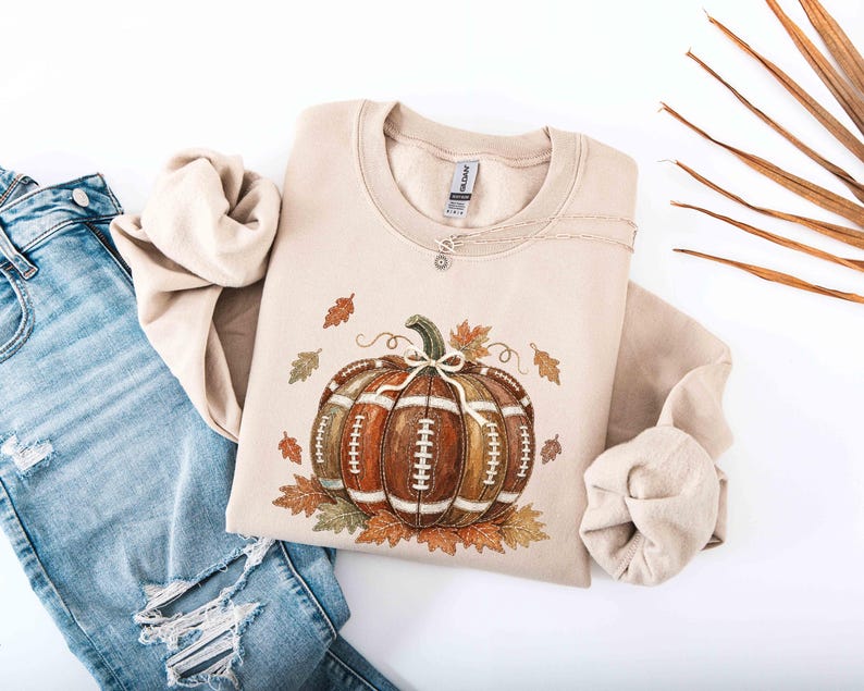 May include: A sand-colored crewneck sweatshirt featuring a graphic of a pumpkin designed with the texture and white laces of an American football, surrounded by various autumn leaves in shades of orange, brown, and green, paired with blue jeans.