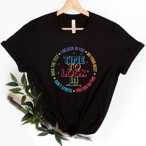 May include: Black t-shirt with a colorful circular design. The design includes the words "TIME TO LOCK IN" in a vibrant font, surrounded by phrases like "Rock the test" and "You can do this."