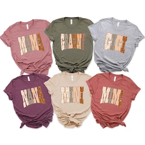 May include: Six short-sleeved crew neck t-shirts in various colors including mauve, olive green, heather gray, plum, beige, and terracotta. Each shirt features a large, arched graphic print with the words MAMA, GRANNY, GIGI, NANA, MINI, or MIMI.