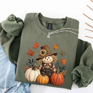 May include: An olive green crewneck sweatshirt featuring a graphic of a scarecrow wearing a hat with a sunflower, dressed in plaid and overalls, surrounded by three pumpkins in white and orange, and scattered autumn leaves in shades of red and gold.