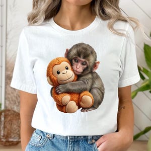 May include: White t-shirt featuring a graphic of two monkeys hugging. One monkey is brown and the other is orange. The design is centered on the shirt.