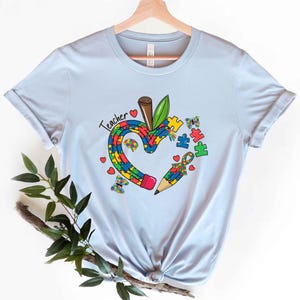 May include: Light blue t-shirt featuring a colorful apple and pencil design with puzzle pieces and butterflies. The word "Teacher" is written above the apple. The apple and pencil are filled with puzzle piece patterns, symbolizing autism awareness.