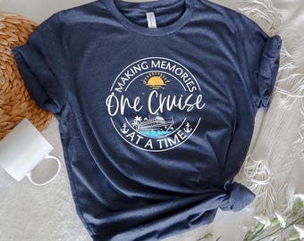 Pode incluir: Camiseta azul marinho com um gráfico circular que diz "Making Memories One Cruise at a Time". O design inclui um navio de cruzeiro, sol e palmeiras. Uma caneca branca e uma cesta tecida estão no fundo.
