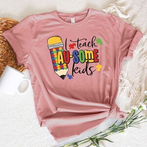 May include: A dusty rose t-shirt with the text "I teach AU-SOME kids." The design features a colorful pencil with puzzle piece details. The shirt is knotted at the bottom and has rolled-up sleeves.