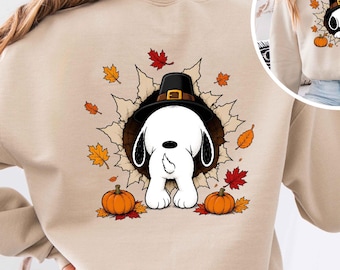 Peut inclure: Un sweat-shirt beige orné d'un motif représentant un chien blanc portant un chapeau de pèlerin noir, sortant d'un trou sombre. Le dessin est entouré de feuilles d'automne orange, jaunes et rouges et de deux citrouilles orange en bas.