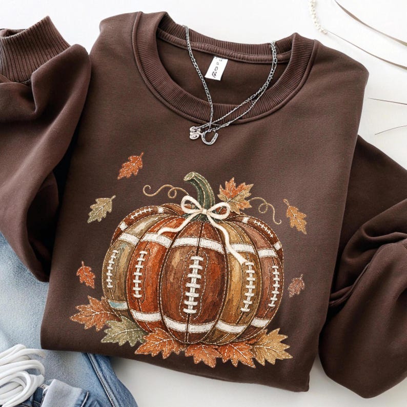 May include: A dark brown crewneck sweatshirt featuring a central graphic of a pumpkin designed with the texture and white laces of an American football. The pumpkin is adorned with a small white bow and surrounded by various autumn-colored leaves.