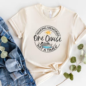 Peut inclure: T-shirt cr&egrave;me &agrave; manches retrouss&eacute;es avec un motif circulaire indiquant "Making Memories One Cruise at a Time". Le motif comprend un bateau de croisi&egrave;re, le soleil, des oiseaux et une ancre. Une veste en jean et de la verdure sont &agrave; c&ocirc;t&eacute; du t-shirt.